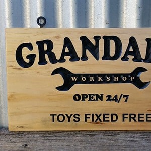Pine sign with carved black engraved lettering and black screw eyes for easy hanging. Reads 'Granddad's workshop open 24/7 toys fixed free'