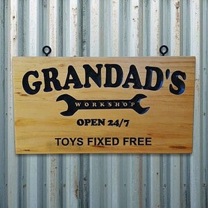 Engraved pine sign with ‘Grandad’s Workshop’ inside a spanner graphic and the text ‘Open 24/7 Toys Fixed Free’ hanging on a corrugated iron shed wall.