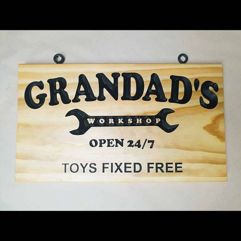 Engraved pine sign with ‘Grandad’s Workshop’ inside a spanner graphic and the text ‘Open 24/7 Toys Fixed Free’ with black screw eyes. Laying flat on the table.