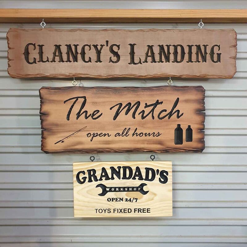Engraved pine sign with ‘Grandad’s Workshop’ inside a spanner graphic and the text ‘Open 24/7 Toys Fixed Free’ hanging from other custom wood signs inside a shed roller door with screw eyes and hooks.