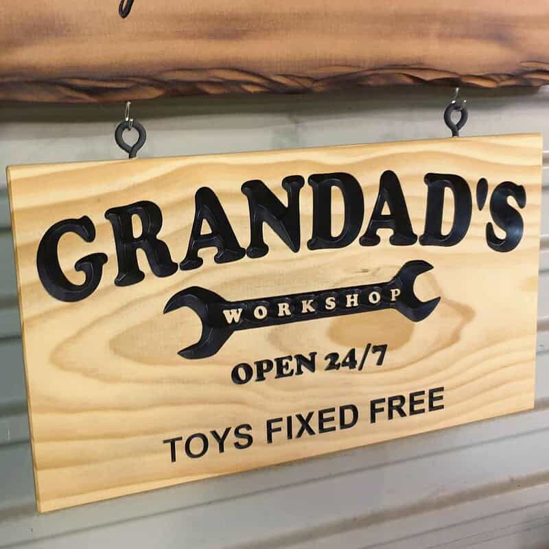 Engraved pine sign with ‘Grandad’s Workshop’ inside a spanner graphic and the text ‘Open 24/7 Toys Fixed Free’ hanging inside a shed roller door from screw eyes and hooks.