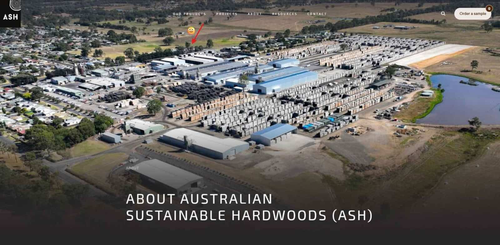 Aerial view of a large sustainable timber processing yard with rows of stacked logs and blue warehouse buildings amid a rural landscape at the edge of fields and a pond nearby. Showing the location of ausworkshop with a red arrow and smiley face.