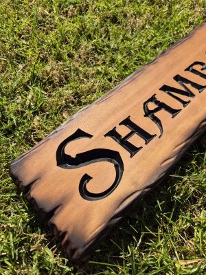 Weathered wooden sign lying on grass with the house name SHAMBAANI carved in bold lettering.