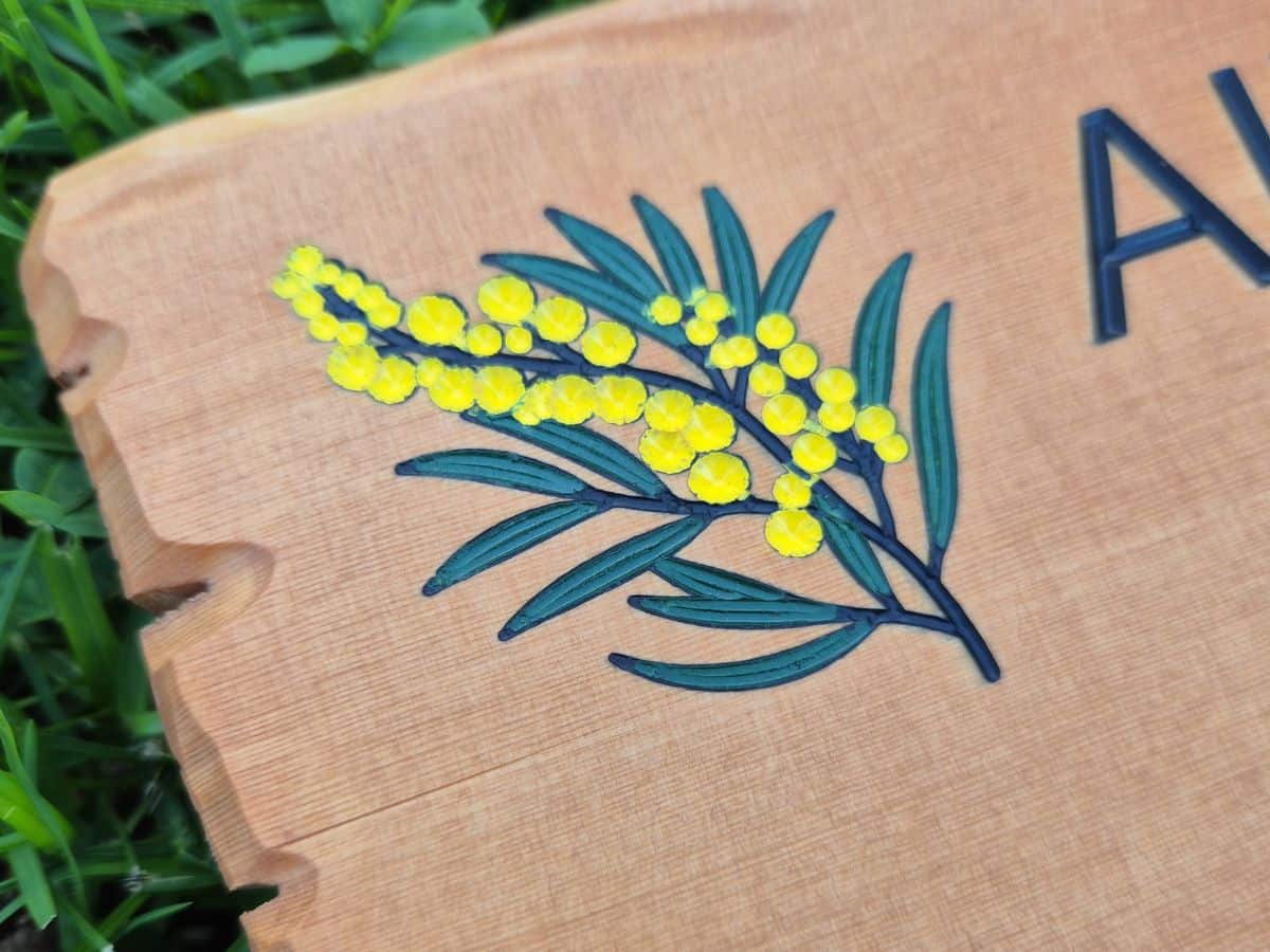 Carved wooden sign with a painted wattle design featuring yellow flowers and green leaves.