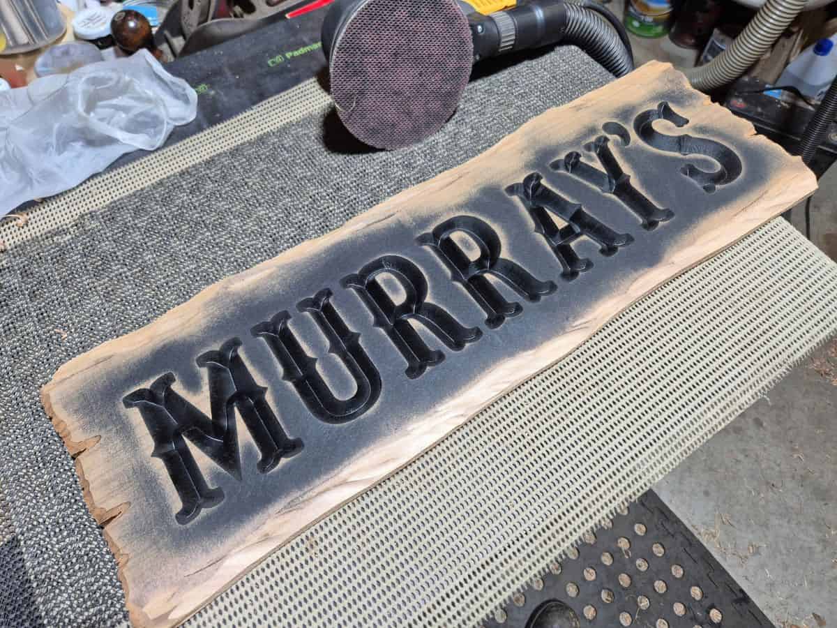 Carved wooden sign with black painted lettering and overspray starting to be sanded from the timber surface.