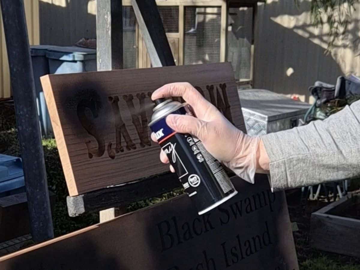 Applying black spray paint to carved lettering on a wooden sign using light controlled passes.